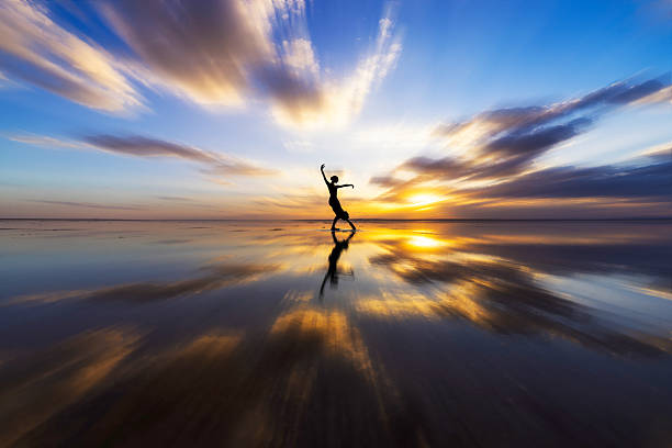 A solitary dancer performs gracefully on a reflective water surface at sunset. The silhouette creates a poetic balance between sky and reflection, symbolizing freedom, movement, harmony, and inner expression. Minimalist composition with dramatic clouds and warm golden light.