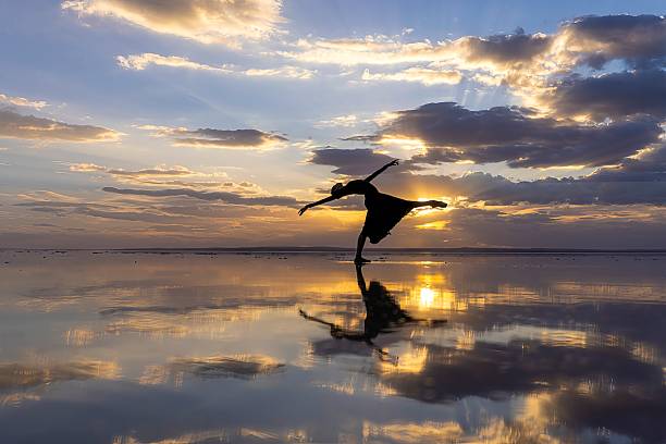 A solitary dancer performs gracefully on a reflective water surface at sunset. The silhouette creates a poetic balance between sky and reflection, symbolizing freedom, movement, harmony, and inner expression. Minimalist composition with dramatic clouds and warm golden light.