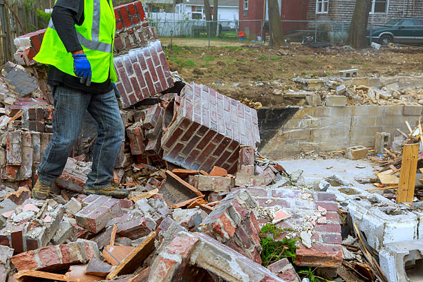 A significant amount of debris lies scattered on the ground where a structure once stood. Surrounding plants and homes are visible, showing signs of recent construction activity.