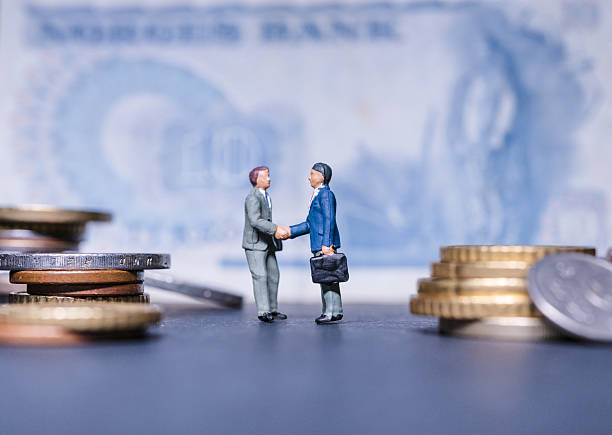 istock Miniature executives shake hands among scattered coins and old banknote. 2251775000