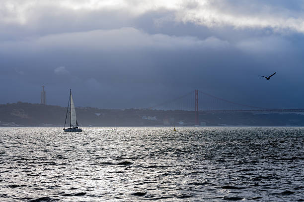 Lisbon Portugal December 05 2025. A sailboat moves along the Tagus River with the Cristo Rei monument visible under heavy winter clouds.