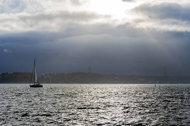 Lisbon Portugal December 05 2025. A sailboat travels across the Tagus River with the Cristo Rei monument and the 25 de Abril Bridge visible under cloudy winter light.