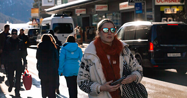 Davos, Switzerland – January 21, 2025: A woman wearing winter clothing walks along a city street in Davos while holding a smartphone, with cars, pedestrians, snow, and alpine surroundings visible.