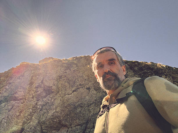 Mature man in his 50s with beard, gray hair & backpack taking selfie during sunny outdoor adventure in the mountains, enjoying nature. Self portrait
