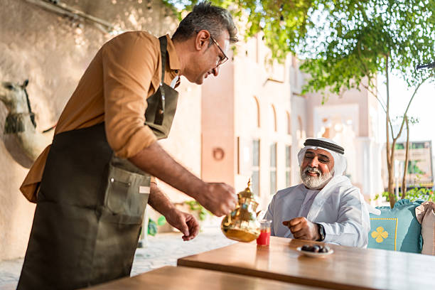 Candid portrait of mid-adult Middle Eastern cafe proprietor in brown jacket and apron serving traditional coffee to elderly gentleman wearing traditional white thobe at outdoor table.