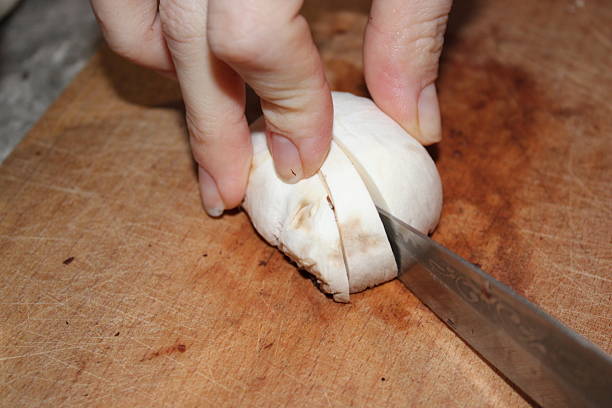 A hand holds a mushroom on a wooden cutting board while using a knife to slice it into pieces. The kitchen environment is visible in the background during meal preparation.
