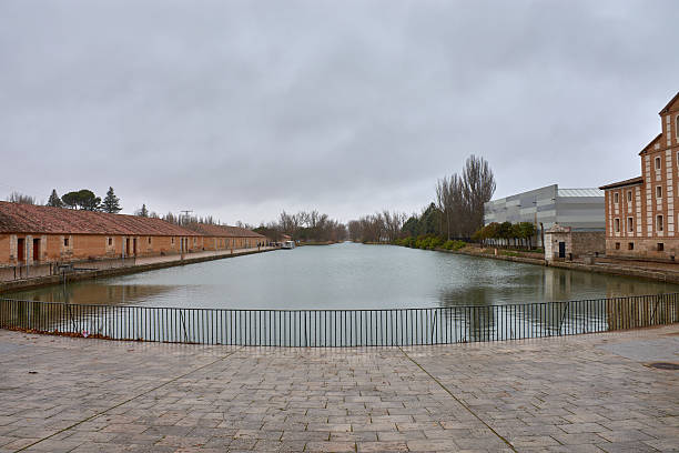 The Castilla Canal Dock in Medina de Rioseco stretches between brick buildings and industrial structures, with a boat docked on the left bank. The gray sky and leafless trees reinforce the winter atmosphere, while the calm canal provides symmetry and serenity. Ideal for content on hydraulic heritage, historical urban planning, cultural tourism, and visual documentation