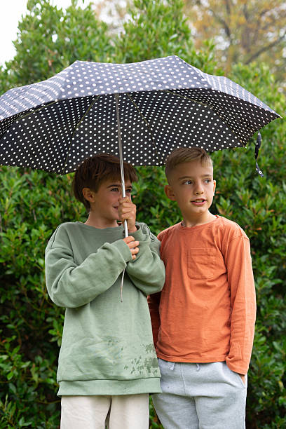istock Two boys sharing umbrella on a rainy day in a park 2251825844