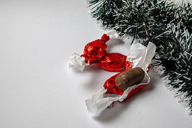 Close-up of wrapped Christmas candies in red foil placed next to a green tinsel decoration on a white background. One candy is partially unwrapped, revealing chocolate inside, creating a festive holiday atmosphere with simple seasonal styling and ample copy space.