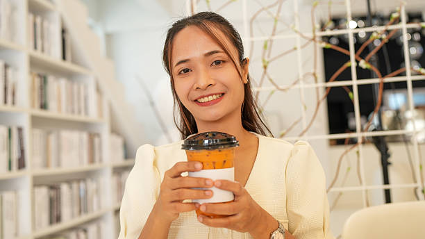 Young woman smiles while holding cup of iced beverage in modern, stylish cafe. bright interior features bookshelves and decorative elements, creating warm and inviting atmosphere