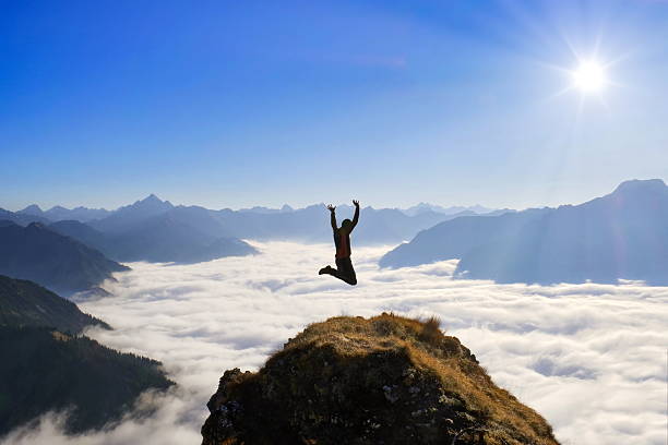 Mountain hiking, man / person jumping on summit with panorama view over landscape, peaks, alps and a foggy dale.