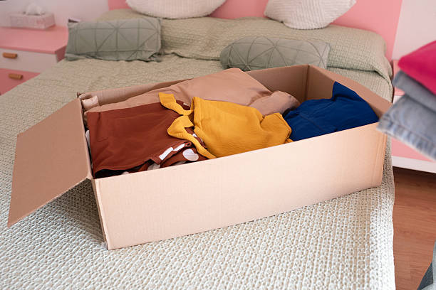 istock Cardboard box filled with assorted clothing items in various colors, placed on a neatly made bed with textured bedding, representing organization and seasonal change 2251832132