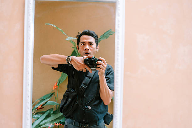 A surprised man takes a self-portrait in a decorative mirror, holding a camera. His wide-eyed expression conveys shock as green plants are reflected in the background, creating a candid moment.
