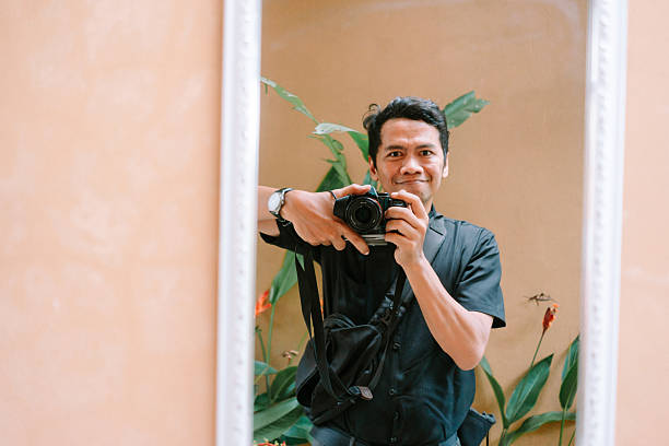 A smiling photographer captures his reflection in a mirror using a digital camera. The warm-toned room features tropical plants in the background, creating a pleasant atmosphere.