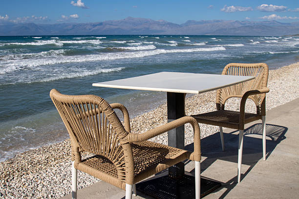 Terrace of a restaurant on the coast of Ionian sea, above the beach. Empty during siesta time. Two chairs and a table. Intense waves. Sunny day. Acharavi, Corfu (Korfu), Greece.