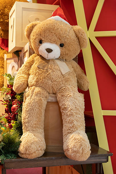 A soft tan teddy bear wearing a red Santa hat sits among festive Christmas decor, gift boxes, and holiday