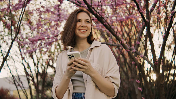 istock Smiling woman stands among cherry blossom trees while using her phone in a garden during the spring season 2251841955