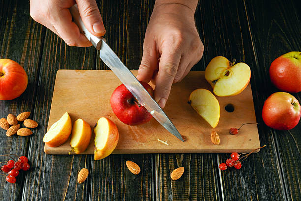 istock Hands expertly slice a red apple on a wooden cutting board, surrounded by cut apple pieces, almonds, and red berries. The scene captures a warm kitchen and active food preparation 2251848698
