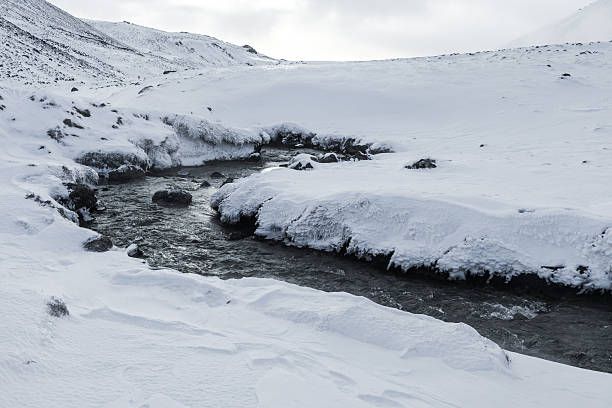 Geothermal hot spring stream flowing through a snow covered Reykjadalur Valley in Iceland, surrounded by winter hills and frozen volcanic landscape.