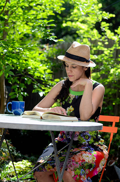 An adult woman is enjoying a quiet moment in a lush garden while reading a book and holding a cup. She sits on an orange chair surrounded by greenery and natural sunlight, creating a peaceful and leisurely outdoor scene. Concept of relaxation, summer lifestyle, self-care, and slow living.