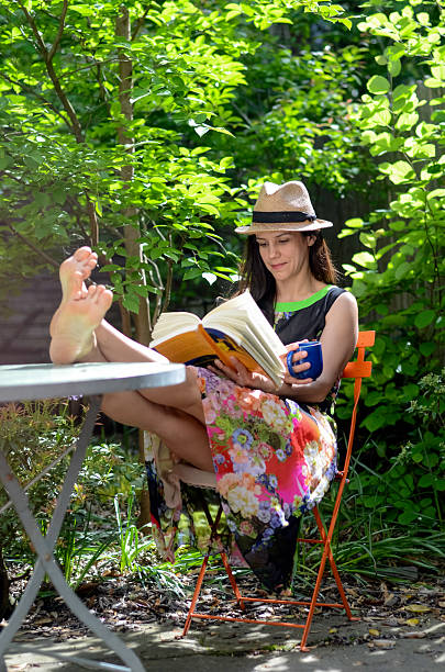 Lady enjoying summer afternoon with book and cold drink in green garden, casual relaxation scene with pet visible behind her.