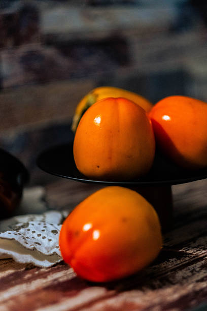 Three persimmons sit on a black plate on a wooden table. The persimmons are ripe and ready to eat