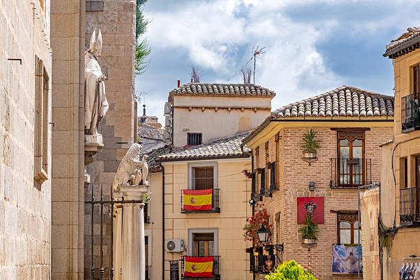 Toledo street corner close-up: Cathedral stone wall with bishop statue and lion contrasts with colorful buildings displaying Spanish flags and wrought-iron balconies