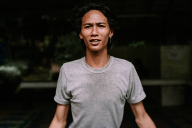 A confident young Asian man with dark hair looks directly at the camera in a casual portrait. He wears a simple grey t-shirt, set against a softly blurred background.
