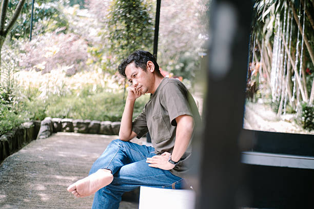 A pensive man sits outdoors, deep in thought, surrounded by the peaceful greenery of a garden or park. He appears relaxed yet contemplative, enjoying a quiet moment in nature.