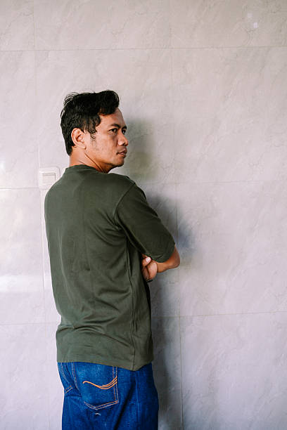 A young man in a casual green t-shirt and jeans stands with arms crossed, looking thoughtfully over his shoulder against a plain tiled wall. The image conveys introspection or observation.