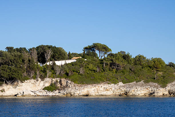 Bright blue water of the Ionian sea. View on a rocky coast, covered by trees and vegetation. Blue sky. Paxos, Greece.