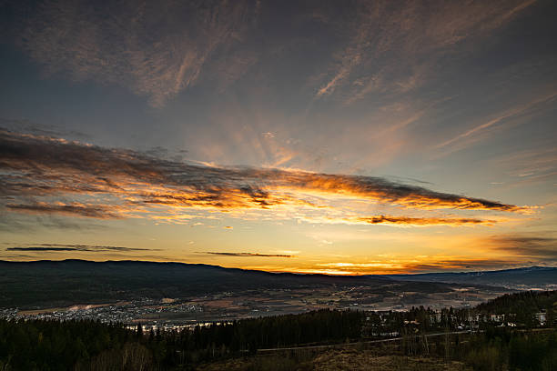 Sun rays spread across the sky above a rural valley during sunset. Warm evening light illuminates layered clouds and distant hills, creating depth and atmosphere over the landscape.