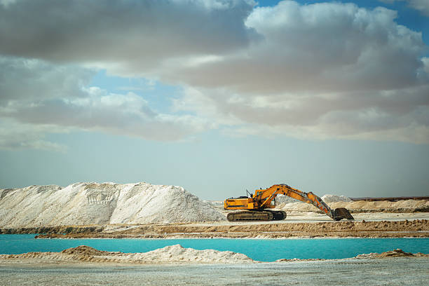 Yellow excavator working in the Siwa salt flat, surrounded by white salt mounds and bright turquoise water under a blue sky with dense clouds.