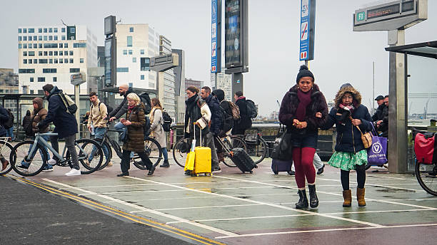 Editorial photograph taken on December 9, 2025, in Amsterdam. The image captures a busy urban scene near the ferry terminal at Amsterdam Centraal Station. Pedestrians and cyclists move through the wet plaza, some carrying luggage or shopping bags. Modern signage and infrastructure indicate public transport connections, including ferry line F3.