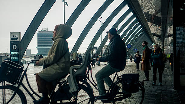 Editorial photograph taken on December 9, 2025, in Amsterdam. The image captures a busy pedestrian and cycling zone near ferry halte D by Amsterdam Centraal Station. People are walking, cycling, and transitioning between transport modes under a modern roof structure with curved beams and reflective surfaces. A cyclist with a child seat, a runner, and several commuters are visible.