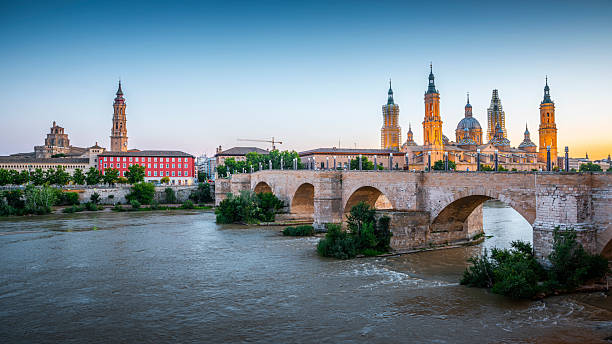Puente de Piedra crossing the Ebro River with the Basilica of Our Lady of the Pillar and Zaragoza skyline in the background. Warm evening light illuminates the historic stone bridge, cathedral towers, and riverbanks. Zaragoza in Aragón, Spain Europe
