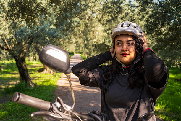 A woman adjusts her helmet beside her bicycle on a peaceful, tree-lined forest path, enjoying a sunny outdoor moment.