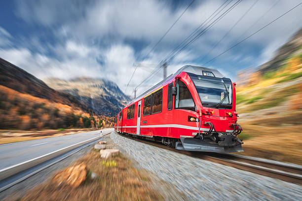 Red modern passenger train is moving on railway in swiss alps at sunset in autumn.  High speed train in mountains, road, orange trees in fall with motion blur effect. Bernina Express, Switzerland