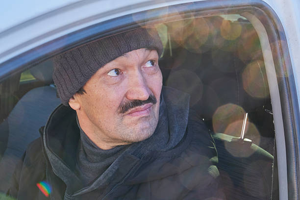 Mature man wearing knitted hat jacket looking intently side car window. Driver seat vehicle cabin waiting for roadside assistance arrival or watching passing event. View from outside in winter day.