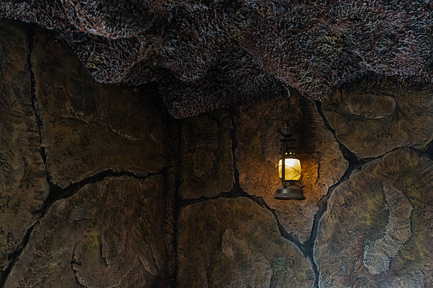A vintage, rustic lantern hangs in a dark cave or grotto, providing warm yellow light that illuminates the surrounding textured rock walls