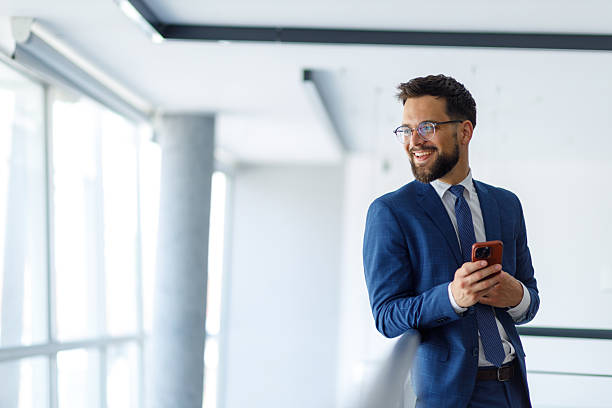 istock Happy young businessman smiling, leaning on railing and using smartphone in modern corporate office 2251910421