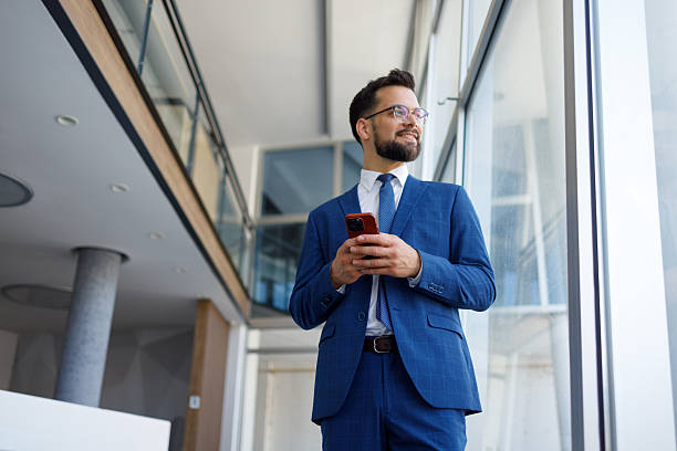 istock Businessman in modern office using mobile phone, smiling and looking out window, thinking about future success 2251911891