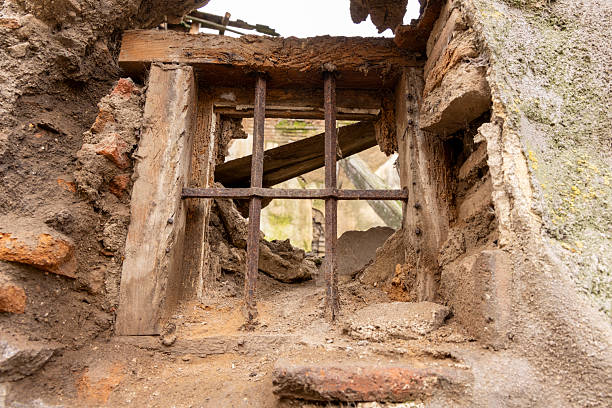 A window with bars on it is shown in a crumbling building. The bars are rusted and the window is small. Scene is one of decay and abandonment
