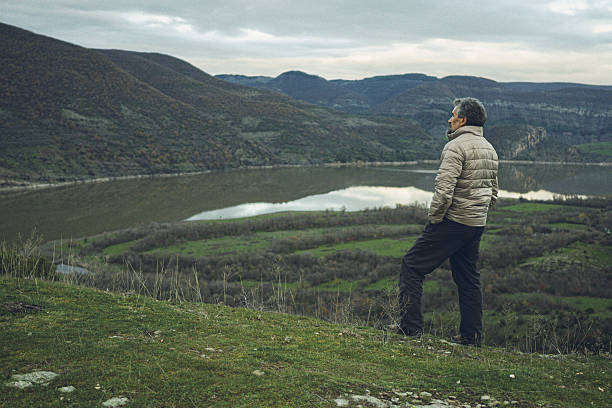 Relaxing mature adult man standing on a hill, overlooking a serene lake and verdant mountains under a cloudy sky, enjoying nature in silence and peace. Serenity