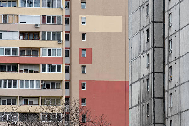 A residential building exterior displays a mix of windows and multicolored wall panels in beige, red, and gray, creating a patchwork appearance. The muted lighting and repetitive geometric elements convey a straightforward urban housing atmosphere.