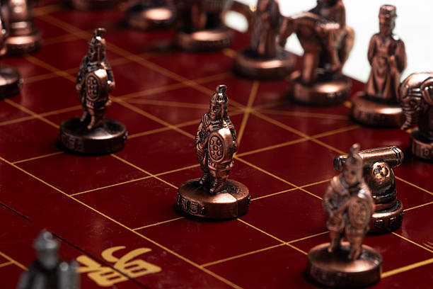 A high-contrast studio photograph of the complete physical Chinese Chess set. The brass and copper pieces evoke themes of competition, heritage, ancient warfare, and critical decision-making processes.