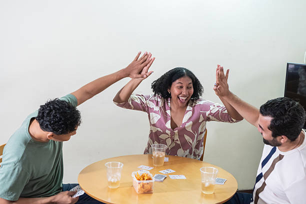 Happy man cheering after winning card game with family at home. Father is celebrating while sitting at table. He is raising his hands.