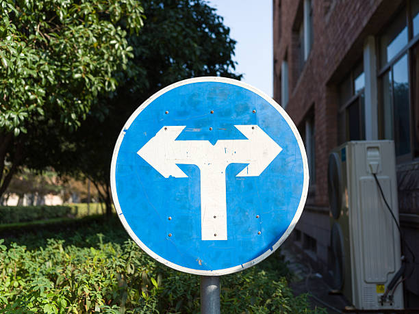 istock Blue sign with T-shaped arrows, building and greenery nearby. 2251928603