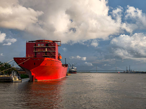 A large red ship is docked in a harbor. The ship is surrounded by water and has a few trees in the background. The scene gives off a sense of calm and tranquility, as the ship is not in motion