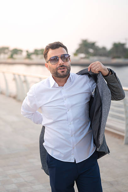 Arab man is adjusting his jacket while wearing sunglasses, exuding casual sophistication by the waterfront, highlighting a stylish demeanor in a serene outdoor environment. Abu Dhabi after lifestyle after working hours